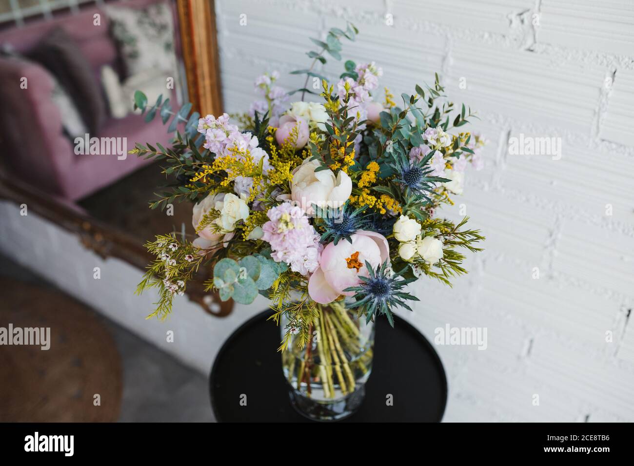 Hoher Winkel von schönen Frühling Bouquet mit verschiedenen bunten blühenden Blumen einschließlich rosa Pfingstrosen und blauen Meer Stechpalme Blumen mit mimosa Zweige auf Glasvase auf Tisch in der Nähe weiß angeordnet Wand Stockfoto