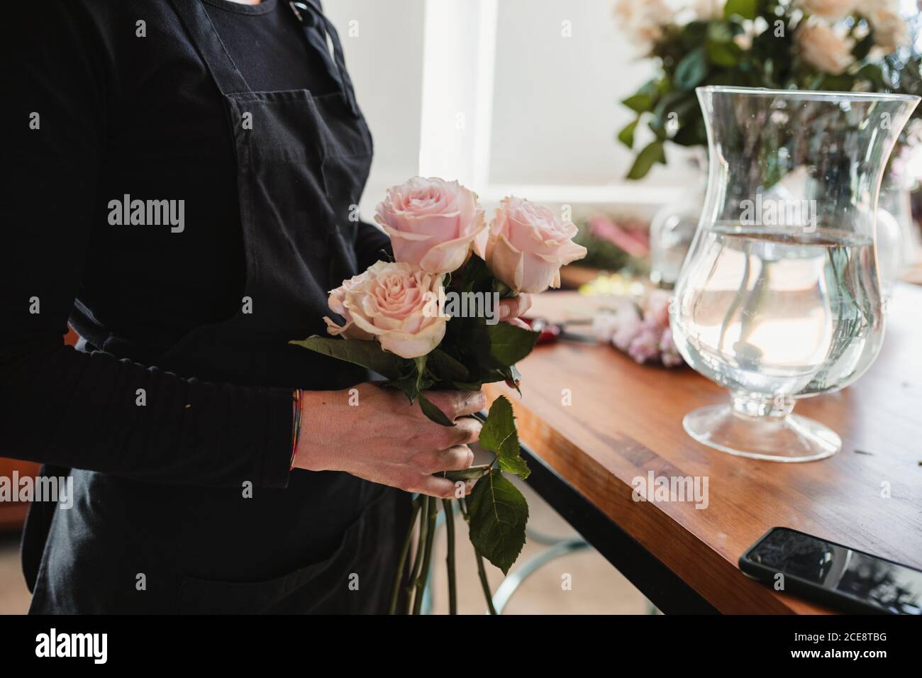 Seitenansicht der Ernte unkenntlich weibliche Floristin hält schön sanft Rosafarbene Rosen beim Stehen an der Theke mit Glasvase gefüllt Mit Wasser während der Arbeit in Floristik Salon Stockfoto