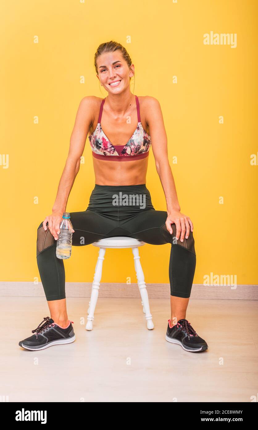 Schlank glücklich weibliche Athletin in Sportkleidung sitzt auf rückenlos Hocker mit Plastikflasche Wasser während der Pause in der Nähe Gelbe Wand und Blick auf die Kamera Stockfoto