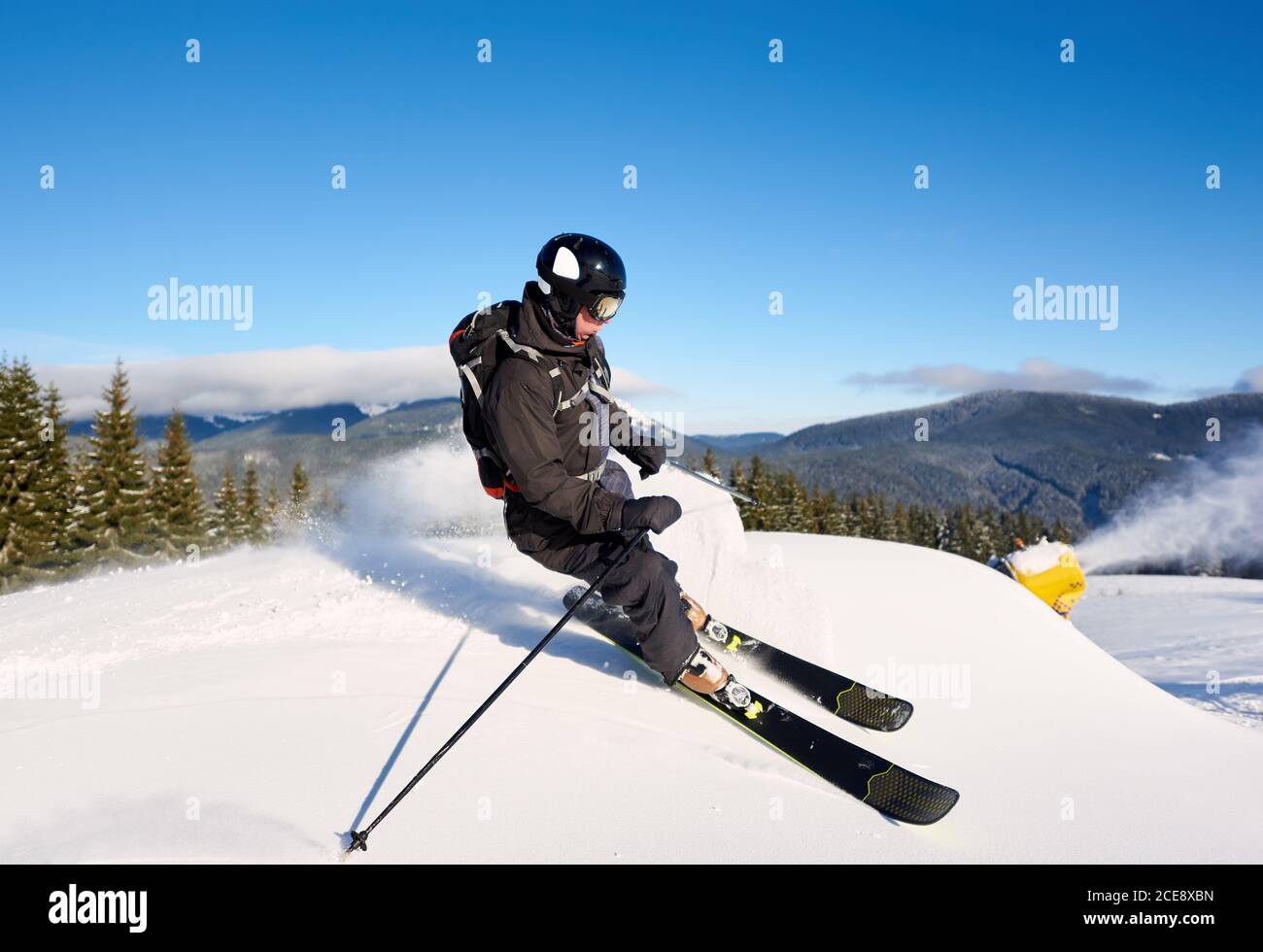 Konzentriertes Mann-Skifahrer-Training auf künstlichem Tiefhang mit frischem Neuschnee an sonnigen Tagen. Elektrische Schneekanone Maschine in Aktion auf Hintergrund. Blauer Himmel und malerische bewaldete Berge im Hintergrund Stockfoto