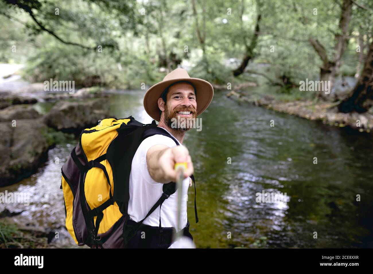Junger Mann mit Barthut auf dem Kopf und gelb Rucksack Wandern auf einer Seenroute mit Bäumen und schattig Bereiche, die ein Selfie machen Stockfoto