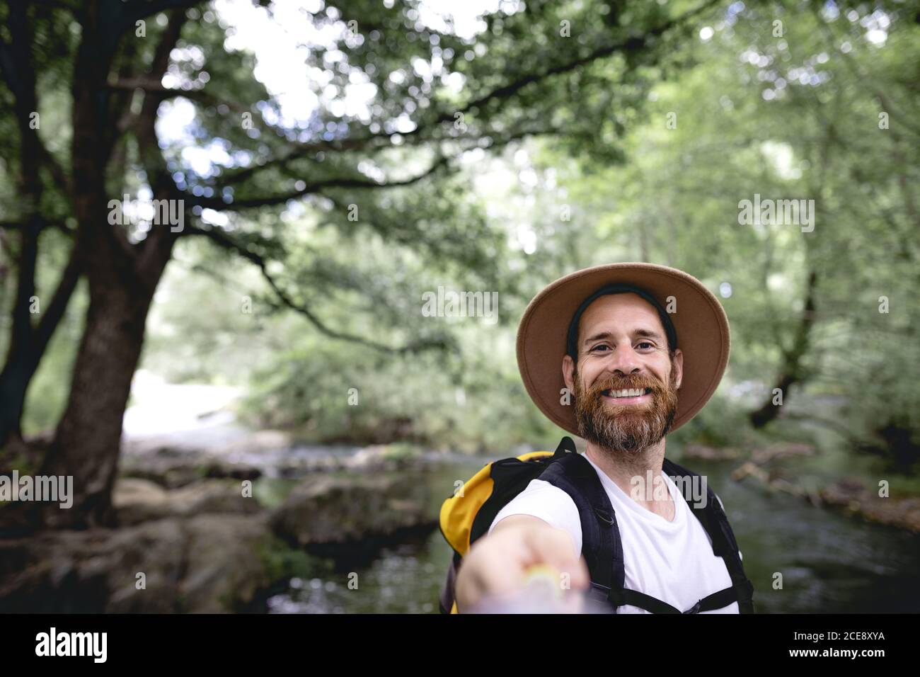 Junger Mann mit Barthut auf dem Kopf und gelb Rucksack Wandern auf einer Seenroute mit Bäumen und schattig Bereiche, die ein Selfie machen Stockfoto