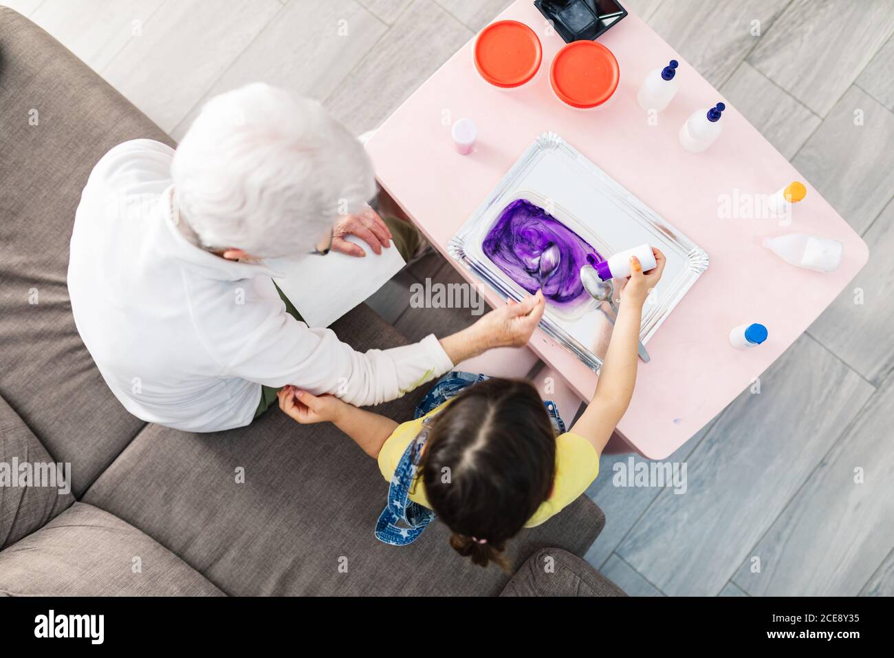 Draufsicht auf grauhaarige Großeltern Rühren bunten Schleim Mischung Mit Löffel in Kunststoff-Behälter, während kleine Mädchen Zugabe von Farbstoff Von Flasche und sitzen auf Sofa in der Wohnung Stockfoto