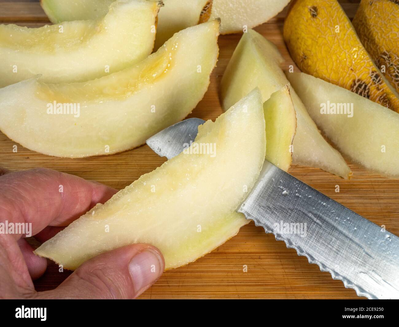 Nahaufnahme eines Mannes Hand hält ein Segment von geschnittenen saftigen Melone, während mit einem scharfen gezackten Stahlmesser, um die äußere Haut / Rinde / Schale zu entfernen. Stockfoto