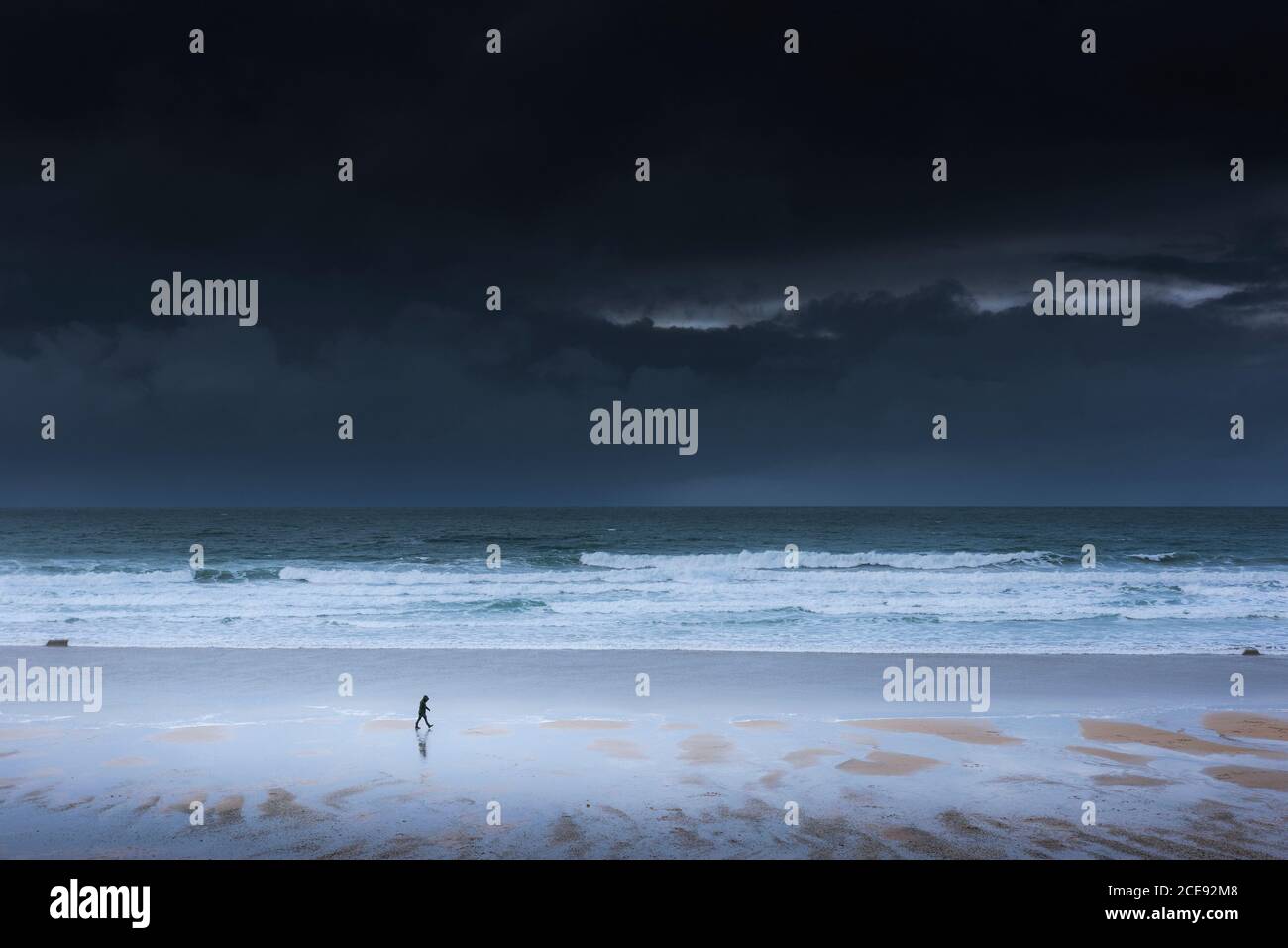 Dunkle Sturmwolken, die sich der Küste nähern, während eine kleine Figur entlang der Küstenlinie von Fistral Beach in Newquay in Cornwall spaziert. Stockfoto