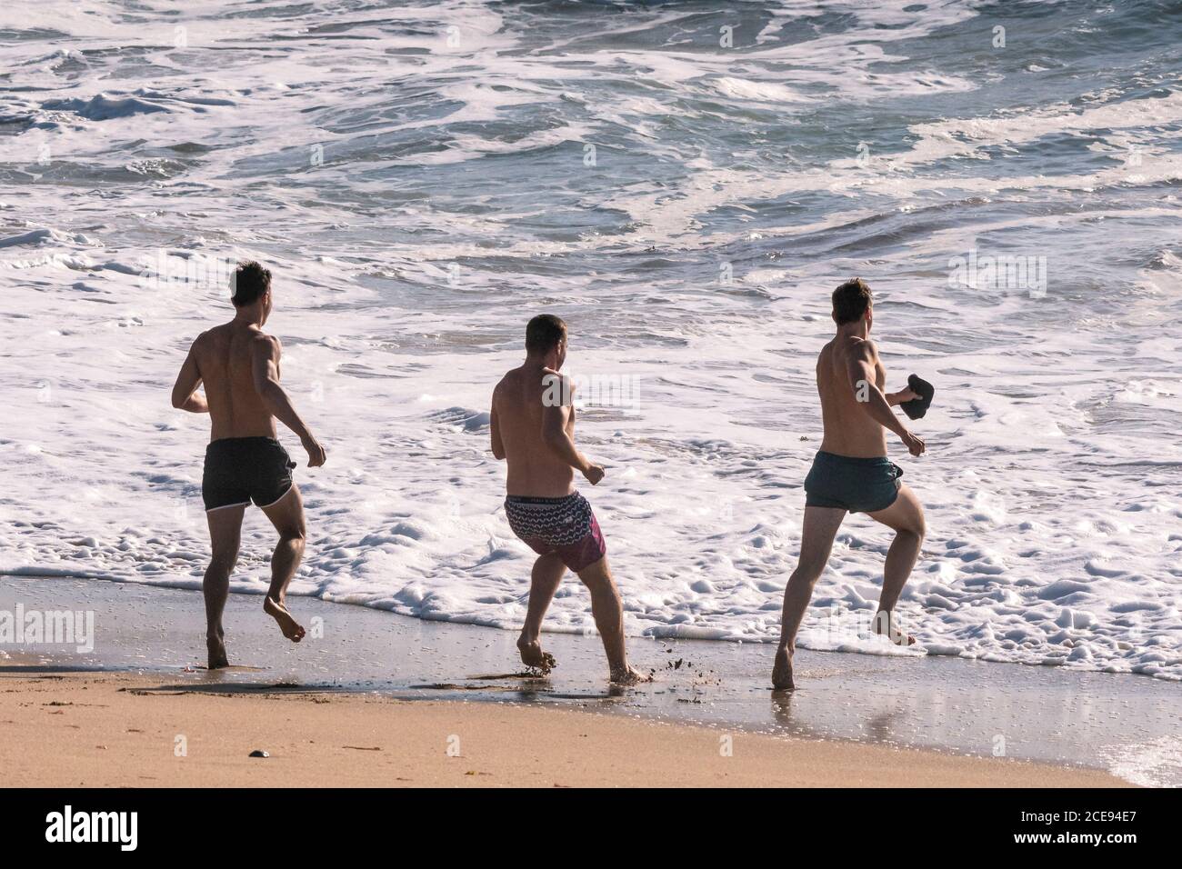 Drei männliche Freunde, die am Fistral Beach in Newquay in Cornwall ins Meer laufen. Stockfoto