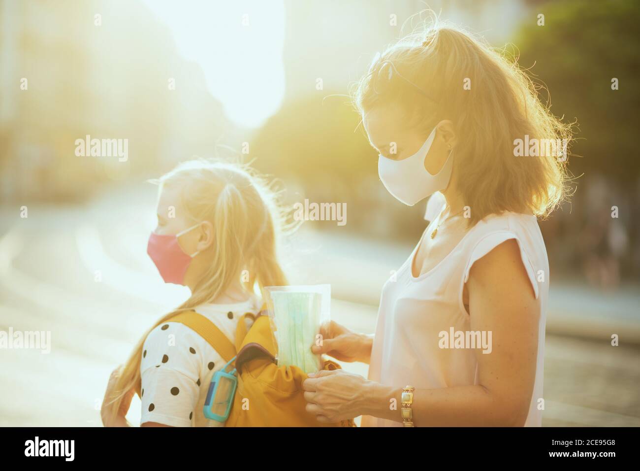 Leben während Coronavirus Pandemie. Junge Mutter und Schulmädchen mit Masken, gelben Rucksack und Desinfektionsmittel Vorbereitung für die Schule und setzen zusätzliche m Stockfoto