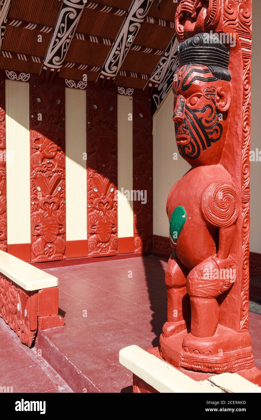 Maori Holzschnitzereien in einem Versammlungshaus in Neuseeland. Die Figur eines Mannes, der eine bloße (Keule) hält, mit dekorativen Wandpaneelen im Hintergrund Stockfoto