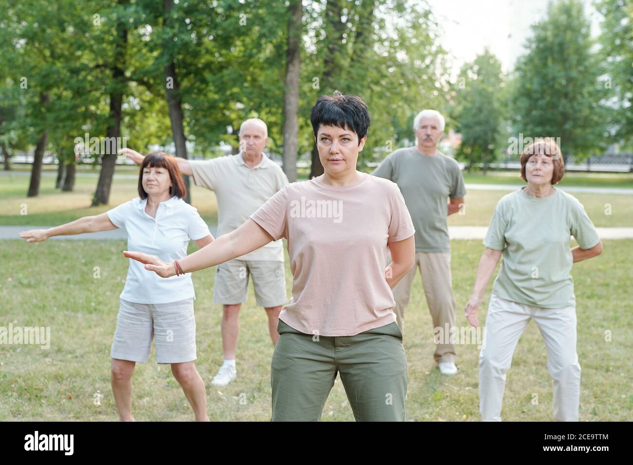 Reifen Brünette Lehrer bewegen Arm beiseite und üben qigong mit Ältere Studenten im Park Stockfoto