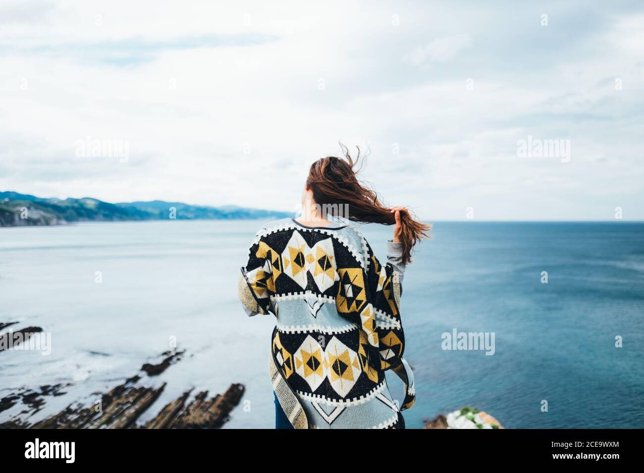 Rückansicht einer Frau mit fliegenden Haaren, die am Rand steht und die Wasserlandschaft bewundert. Stockfoto