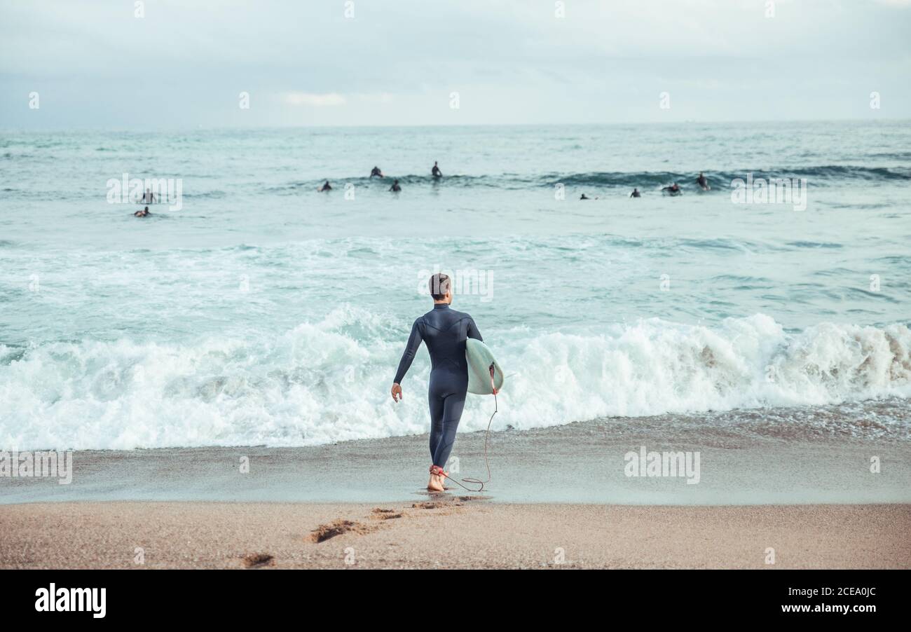 Rückansicht des Mannes im Neoprenanzug, der auf rauhen Wellen des Ozeans mit Surfbrett läuft, Spanien Stockfoto