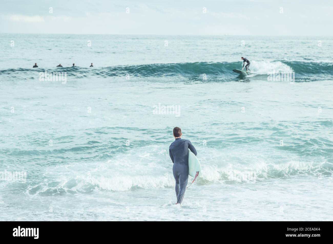 Rückansicht des Mannes im Neoprenanzug, der auf rauhen Wellen des Ozeans mit Surfbrett läuft, Spanien Stockfoto