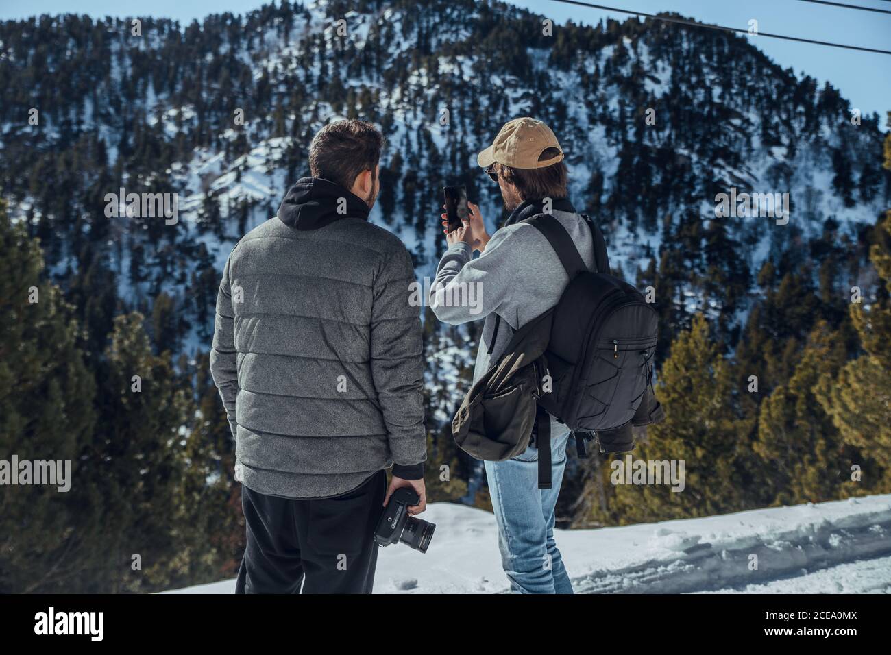 Rückansicht von jungen Fotografen mit professioneller Kamera und Handy stehen zwischen Bergen im Schnee in Cerdanya, Frankreich Stockfoto