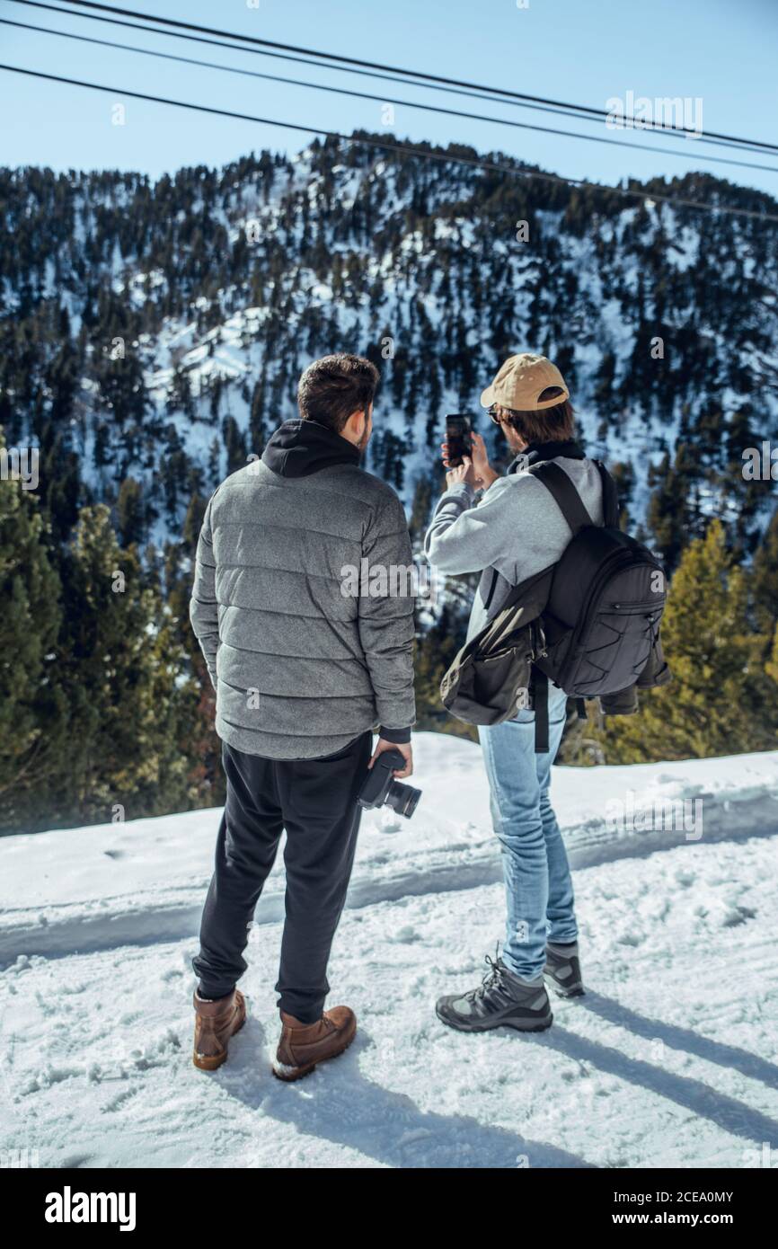 Rückansicht von jungen Fotografen mit professioneller Kamera und Handy stehen zwischen Bergen im Schnee in Cerdanya, Frankreich Stockfoto