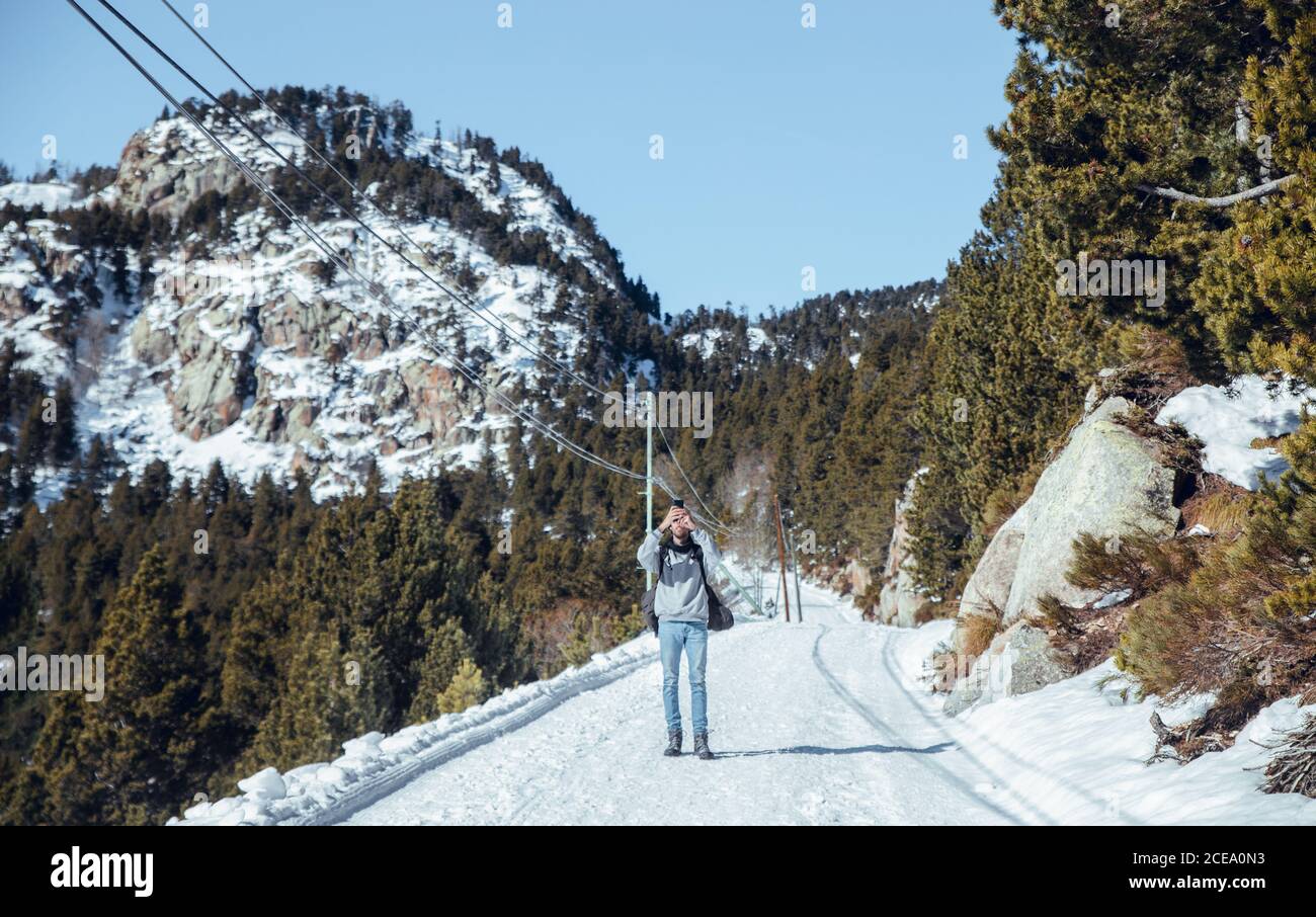 Junger Mann, der mit dem Handy fotografiert und auf der Straße zwischen den Bergen im Schnee in Cerdanya, Frankreich, steht Stockfoto