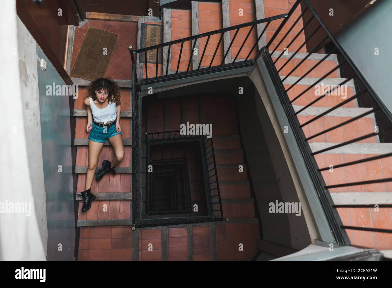 Von oben Aufnahme einer lockigen Frau in Shorts, die auf einer schäbigen Treppe im Flur sitzt und nach oben schaut Stockfoto