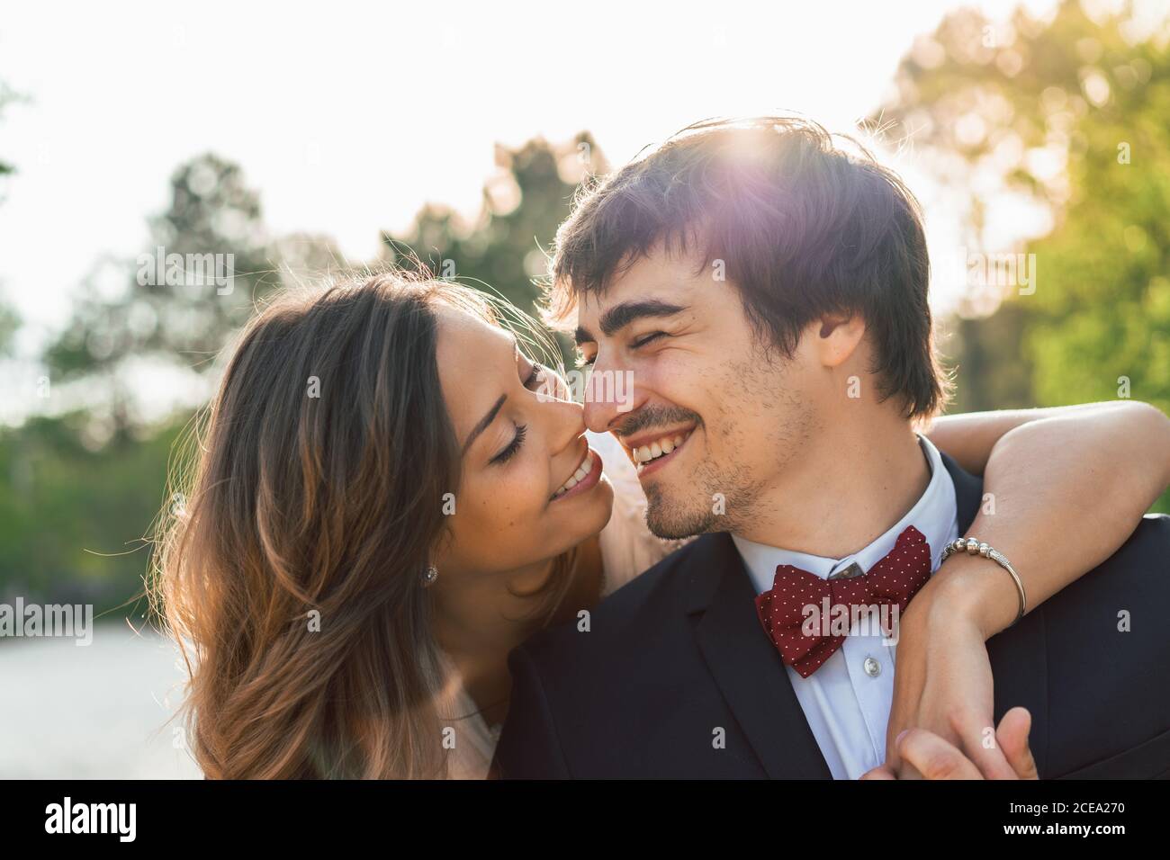 Fröhlicher, eleganter Mann und Frau in Hochzeitsoutfits umarmen sich am Strand und schauen lächelnd im Sonnenlicht weg Stockfoto
