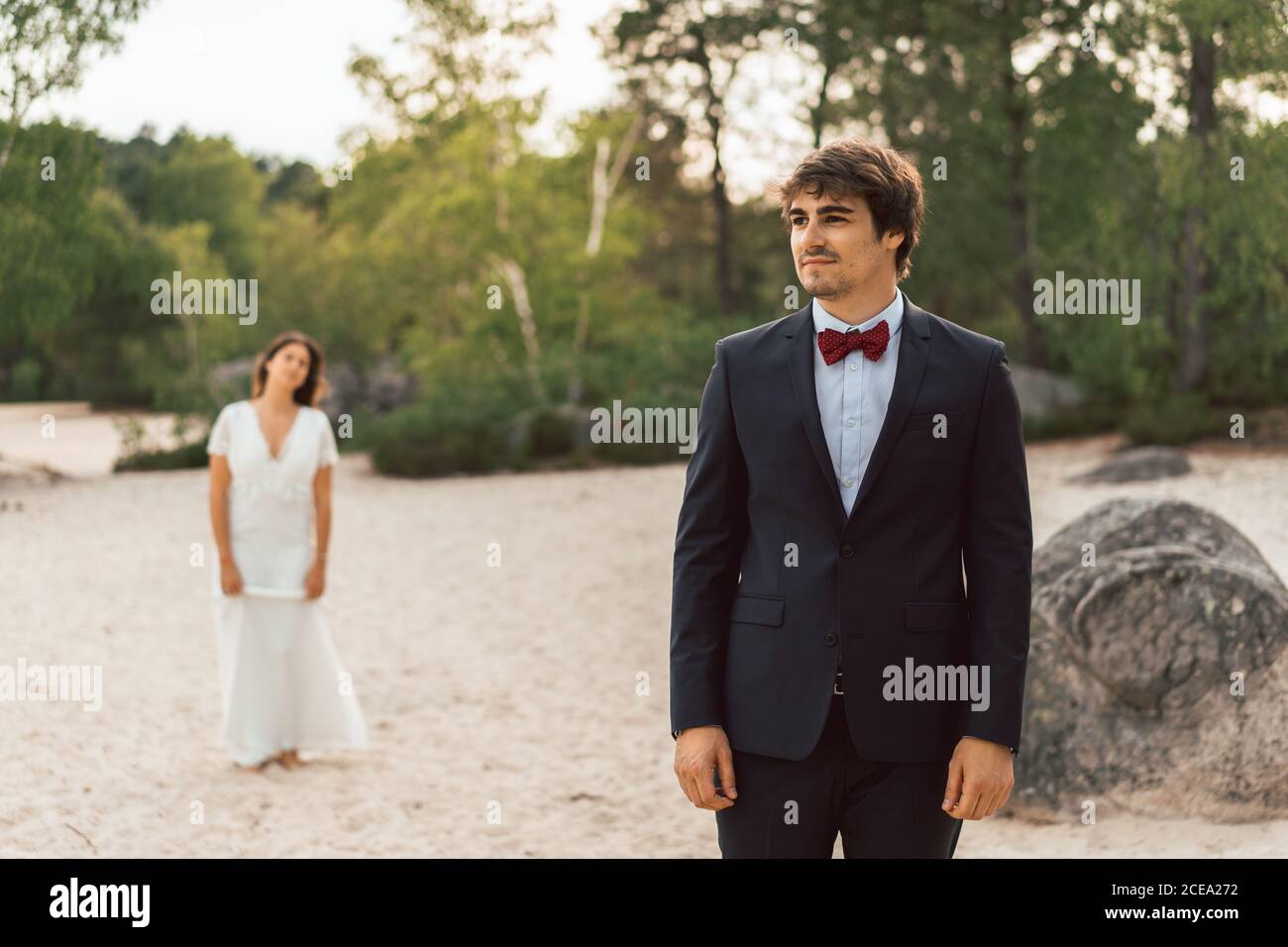 Mann und Frau in eleganten Brautkleidern stehen getrennt auf sandiger Küste mit grünen Bäumen im Sonnenlicht auf dem Hintergrund Stockfoto