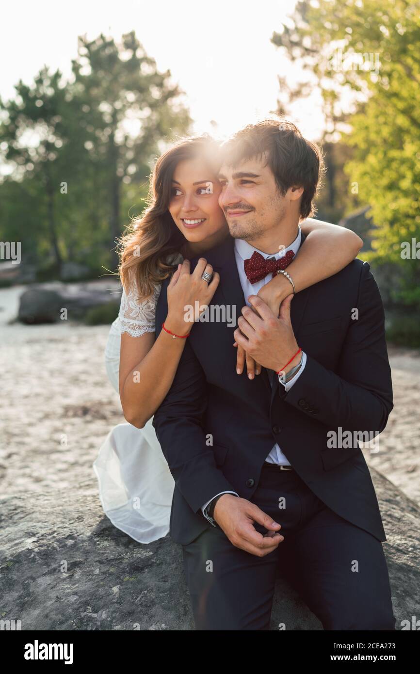 Fröhlicher, eleganter Mann und Frau in Hochzeitsoutfits umarmen sich am Strand und schauen lächelnd im Sonnenlicht weg Stockfoto