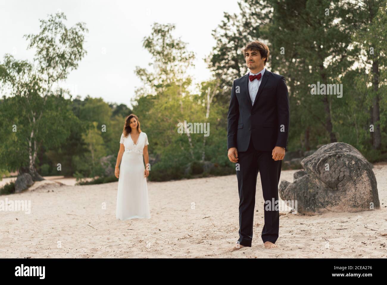 Mann und Frau in eleganten Brautkleidern stehen getrennt auf sandiger Küste mit grünen Bäumen im Sonnenlicht auf dem Hintergrund Stockfoto