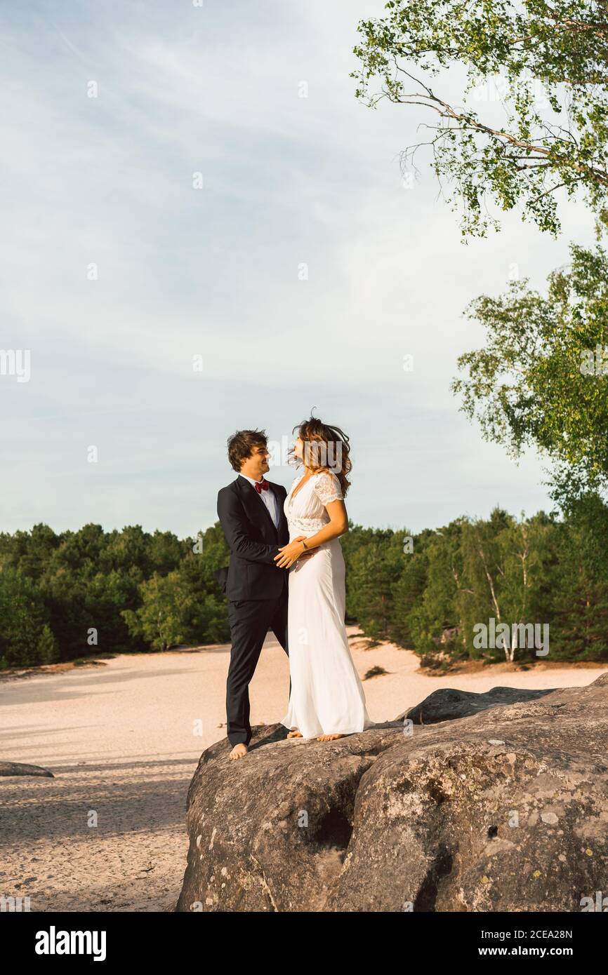 Blick in der Ferne von Paar in Brautkleider stehen auf Felsen und umarmen glücklich gegen grüne Bäume und blauen Himmel Stockfoto