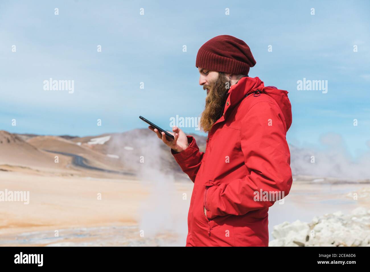 Seitenansicht von bärtigen Mann in warmen hellen Kleidern mit Mobiltelefon, das vor der dampfenden heißen Quelle steht Namaskardh in Island Stockfoto