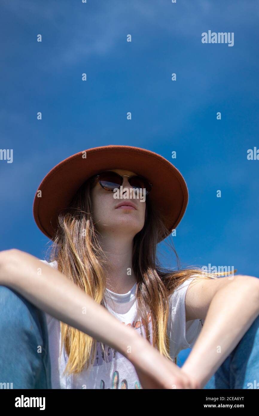 Von unten Aufnahme einer jungen selbstbewussten Frau mit langen Haaren in lässiger Kleidung mit Sonnenbrille und Hut unter blauem Himmel Stockfoto