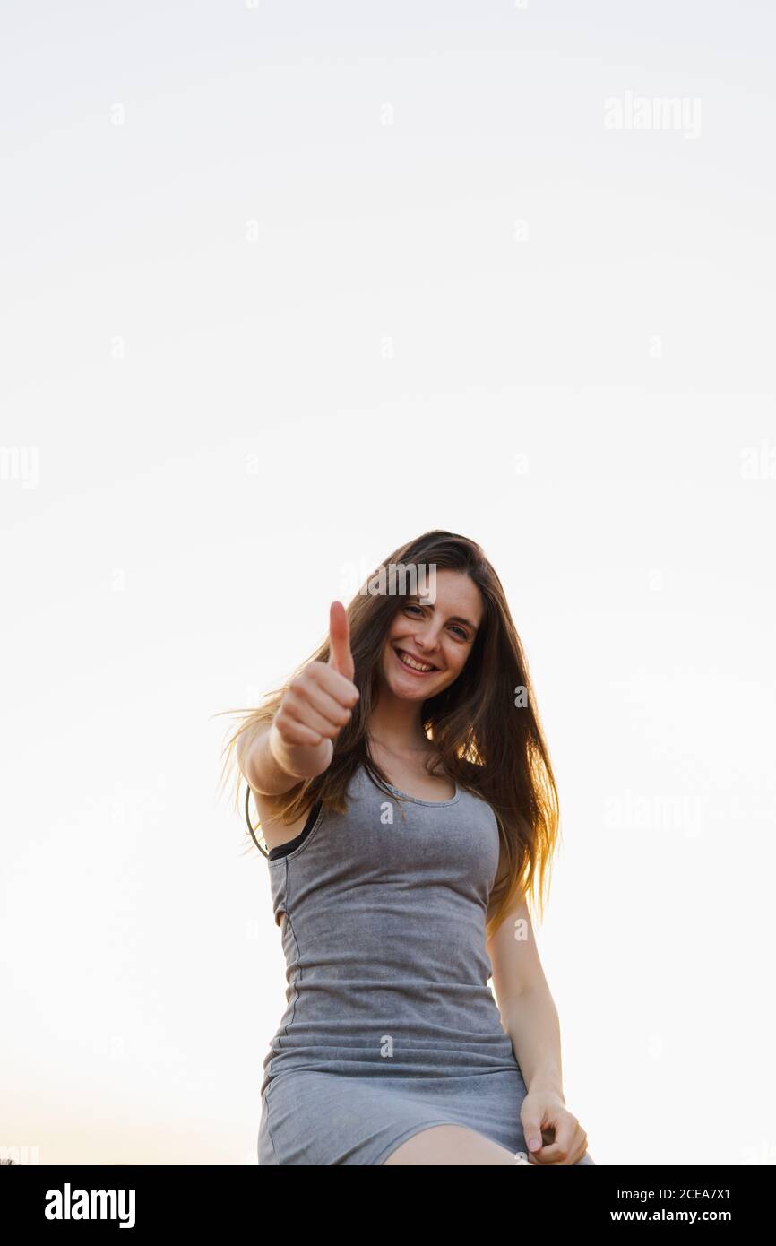 Wunderschöne junge Brünette weiblich stehen im Freien in lässigen Sommerkleid Und zeigt Daumen nach oben Geste Blick auf die Kamera und lächelt Auf hintergrundbeleuchtetem Hintergrund des klaren Himmels von unten Stockfoto