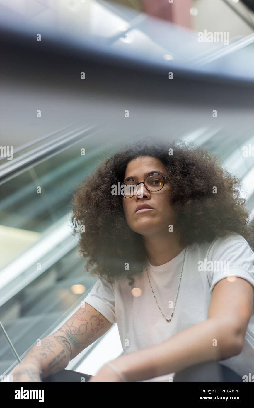 Junger afroamerikanischer Kerl in weißem Hemd und Brille mit Afro Frisur und Tattoos auf der Hand sitzen auf beweglichen Treppe Stockfoto