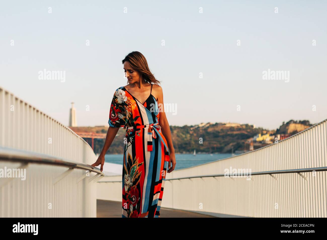 Elegante Frau in Kleid, die sich auf einem Zaun auf einer Brücke lehnt und das Stadtbild im Sonnenlicht beobachtet Stockfoto