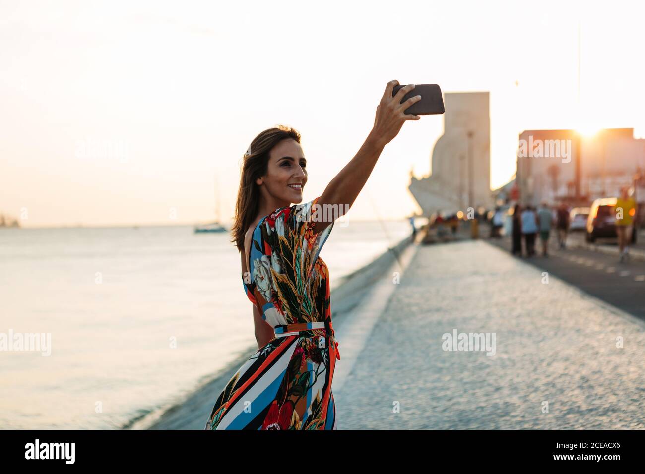Content Frau in Kleid mit Telefon und Selfie auf Strandpromenade bei Sonnenuntergang Stockfoto