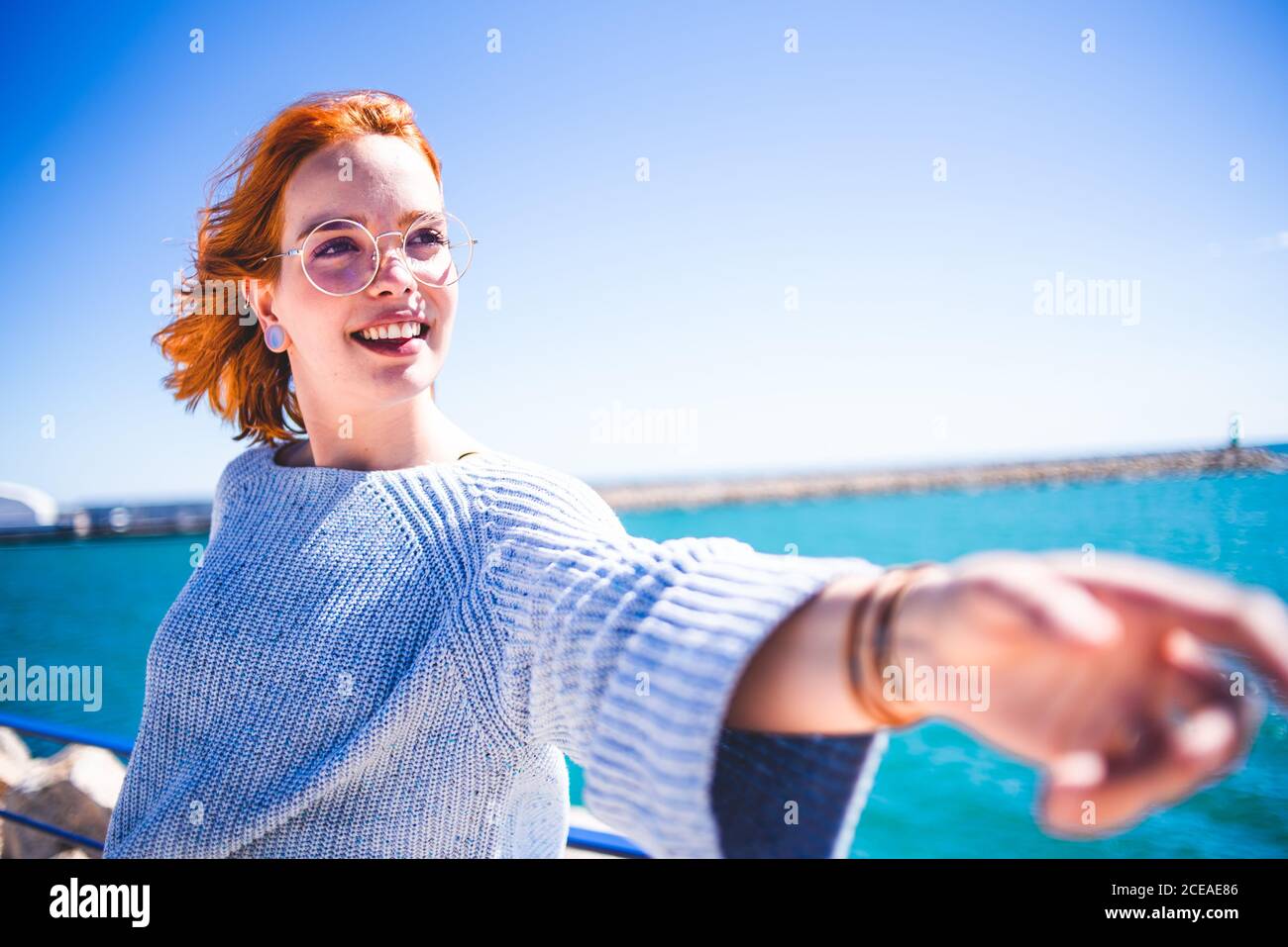 Fröhliche Rothaarige Frau, die an der Hand zieht und wegschaut, während sie sich am Meer auf den Handlauf lehnt. Stockfoto