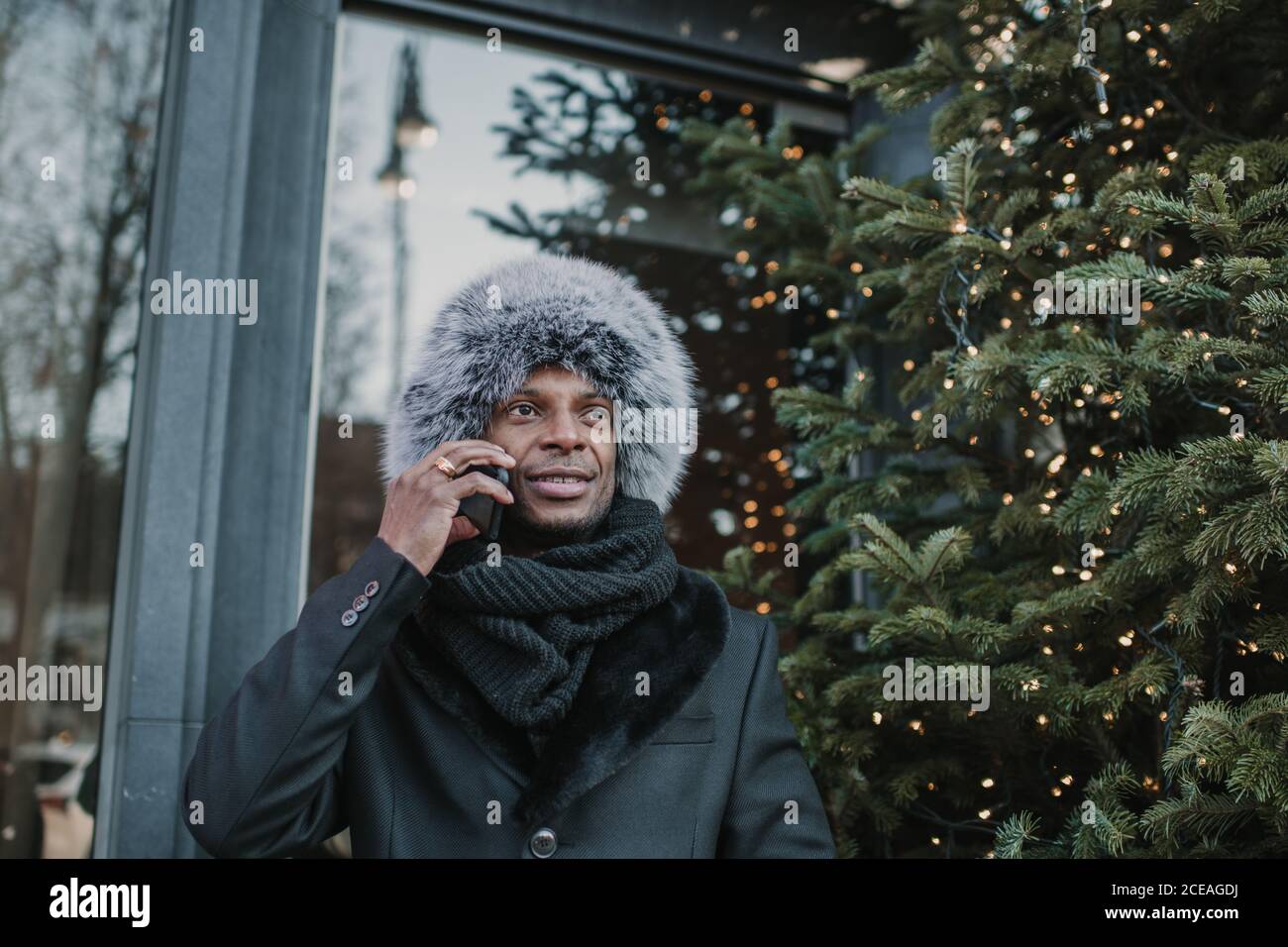 Von unten Schuss aufgeregt afroamerikanischen Männchen in warm Kleidung, die auf dem Smartphone spricht, während sie auf der Stadtstraße in der Nähe steht Bau und Nadelbaum geschmückt für Weihnachten Stockfoto