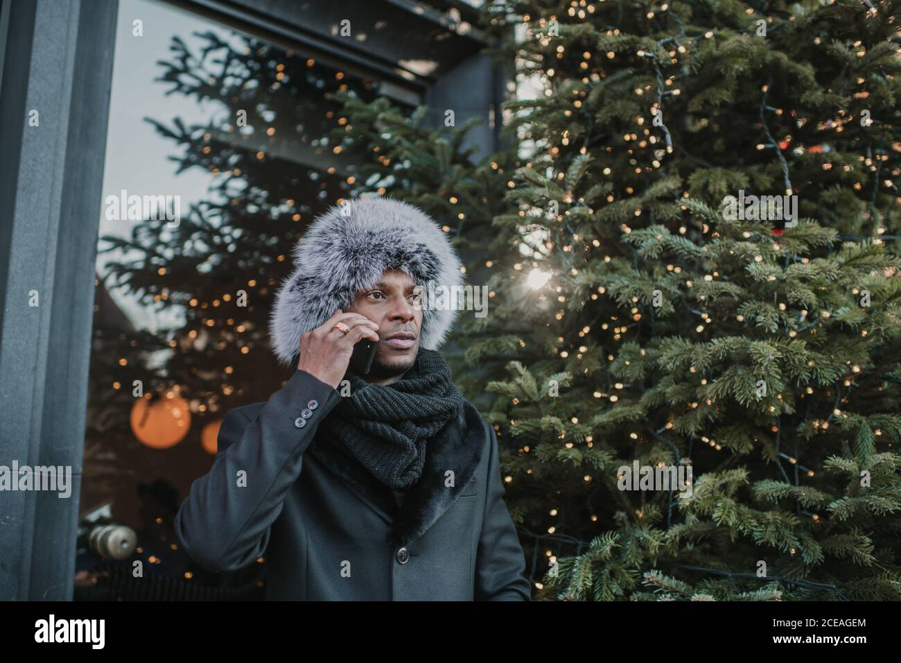Von unten Schuss aufgeregt afroamerikanischen Männchen in warm Kleidung, die auf dem Smartphone spricht, während sie auf der Stadtstraße in der Nähe steht Bau und Nadelbaum geschmückt für Weihnachten Stockfoto