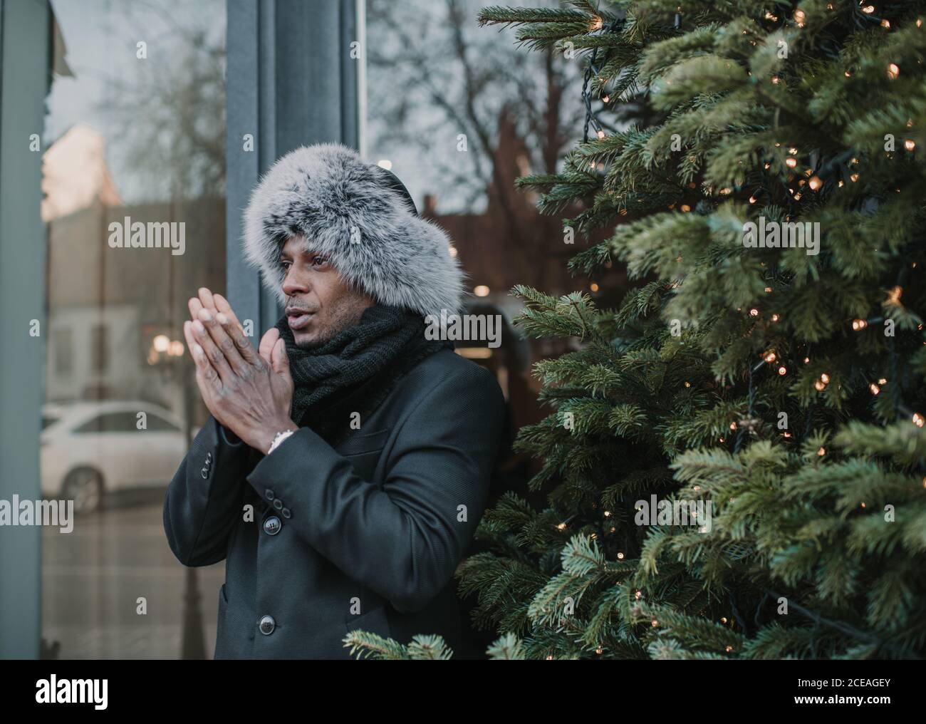Hübscher afroamerikanischer Kerl in warmen Kleidern, der Hände während des Stehens reibt In der Nähe Weihnachtsbaum mit Lichterketten auf der Stadtstraße Stockfoto