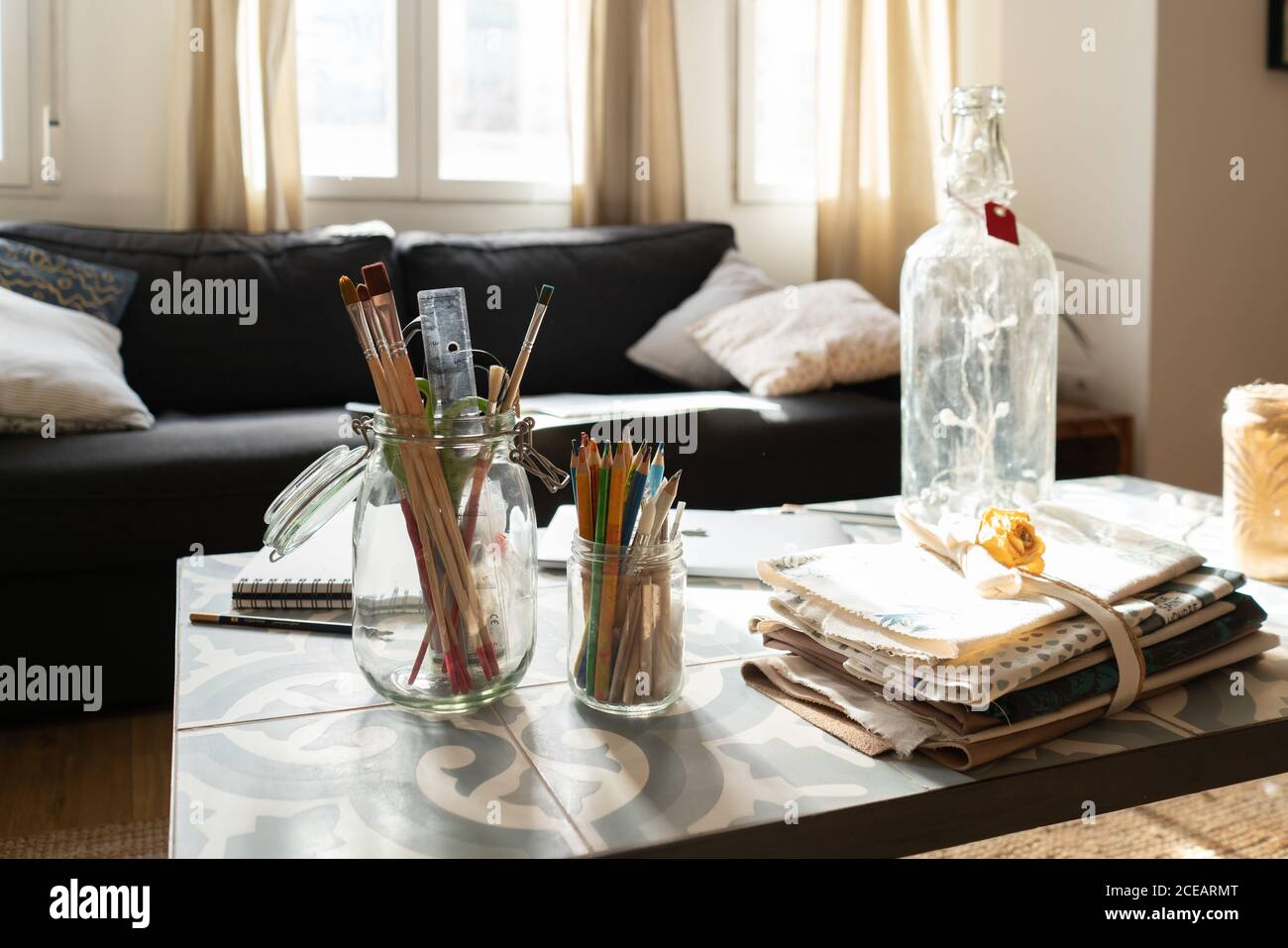 Sammlung von Bürsten und Bleistifte in Glas Dosen in der Nähe von Flasche und Haufen von Materialien auf niedrigen Tisch in der Nähe der Couch im Zimmer Stockfoto