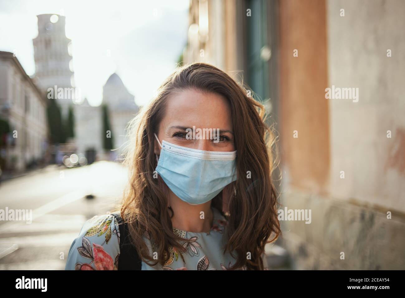 Reisen während der covid-19 Pandemie. Portrait von glücklichen modernen Solo-Tourist Frau in floralen Kleid mit medizinischer Maske in der Nähe schiefen Turm in Pisa, Italien. Stockfoto