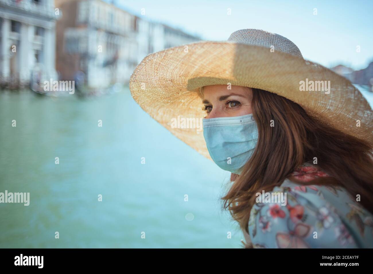 Reise während der Coronavirus-Pandemie. Stilvolle Frau in Blumenkleid mit medizinischer Maske Blick in die Ferne und mit Ausflug auf Vaporetto Wasser Stockfoto