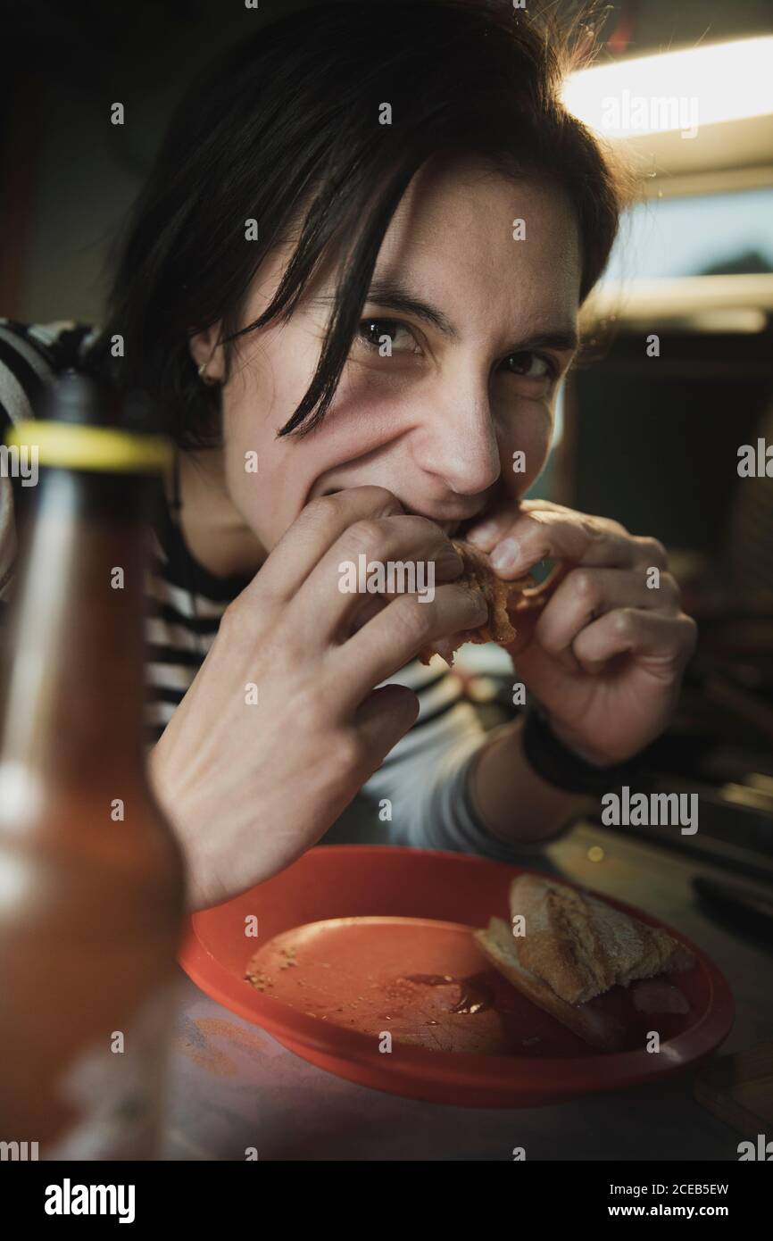 Dame am Tisch, beißen Sandwich in der Nähe von Gericht und eine Flasche trinken und Kamera in Mobile home Stockfoto