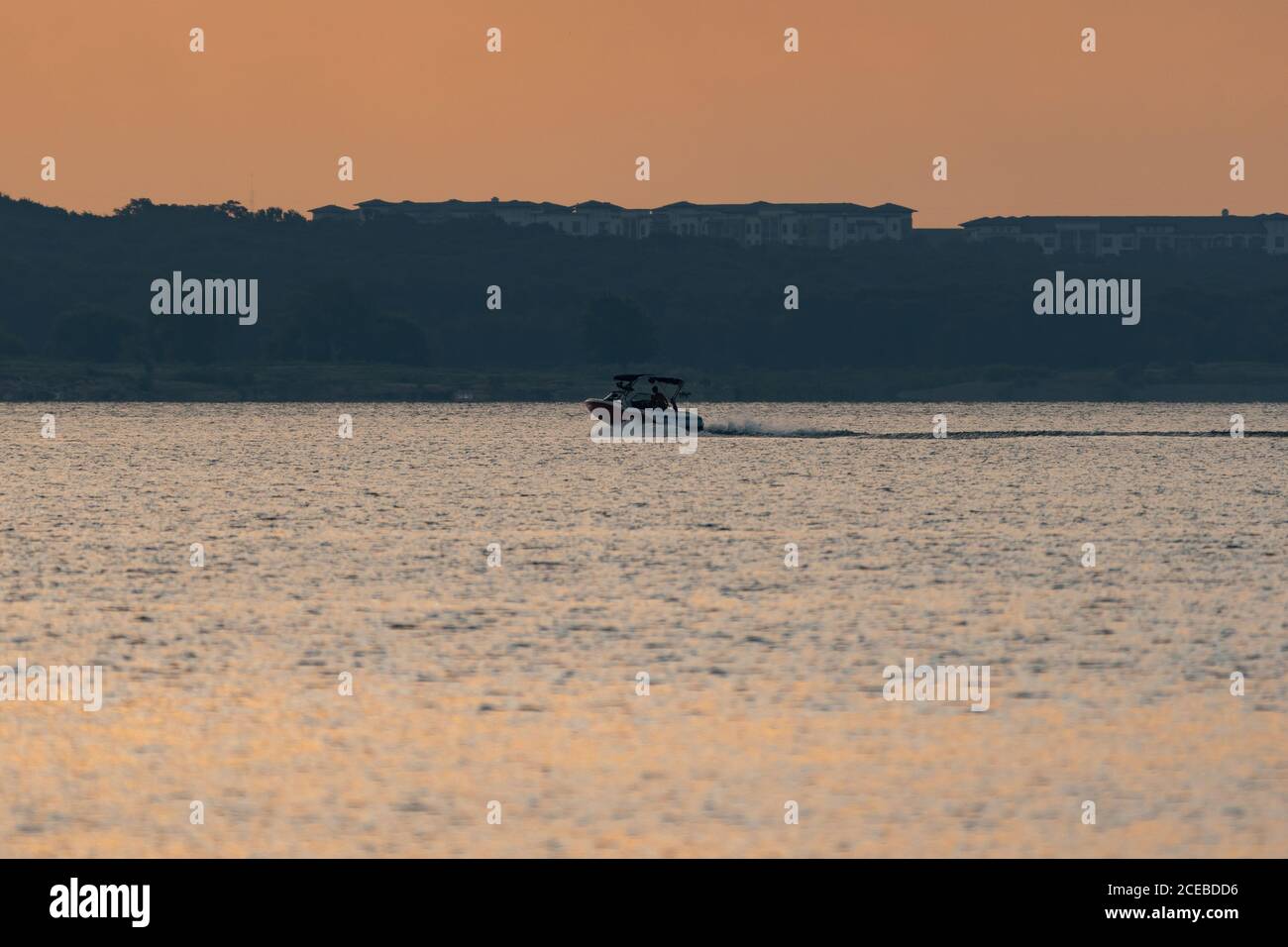 Ein Motorboot, das über einen See rast, kurz nach Sonnenaufgang mit einer hohen Eigentumswohnung, die über den Bäumen am Ufer im Hintergrund aufsteigt. Stockfoto