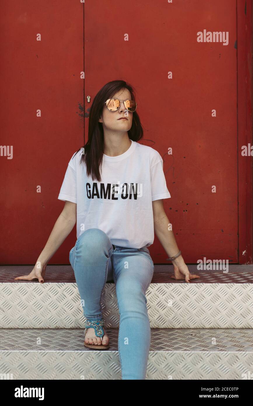 Schöne junge Frau in Sonnenbrille und T-Shirt mit Spiel auf Schreiben wegschauen, während sitzen auf Metalltreppen in der Nähe rot Tür Stockfoto