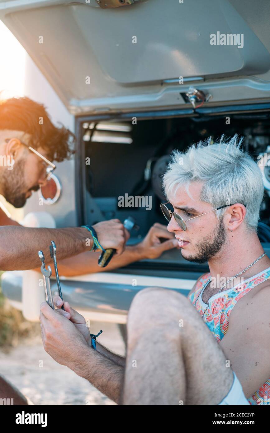 Junger Mann lehnte sich zurück und entspannen, während in der Nähe von Freund sitzen Versuchen Sie, defekten Motor des Autos während der Reise in zu reparieren Natur Stockfoto