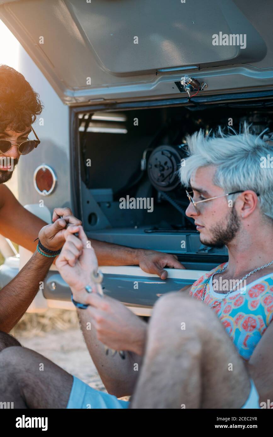 Junger Mann lehnte sich zurück und entspannen, während in der Nähe von Freund sitzen Versuchen Sie, defekten Motor des Autos während der Reise in zu reparieren Natur Stockfoto