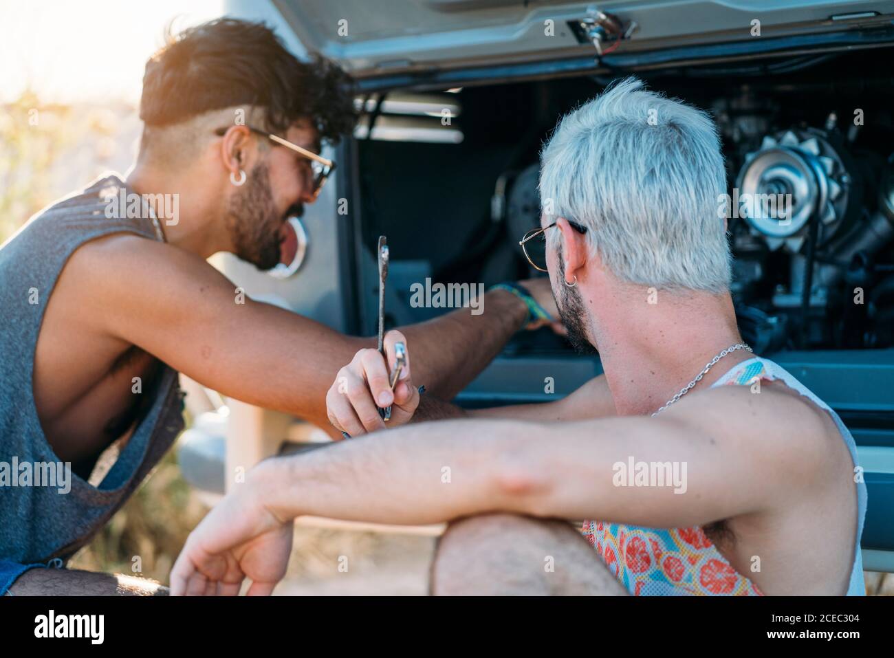 Junger Mann lehnte sich zurück und entspannen, während in der Nähe von Freund sitzen Versuchen Sie, defekten Motor des Autos während der Reise in zu reparieren Natur Stockfoto