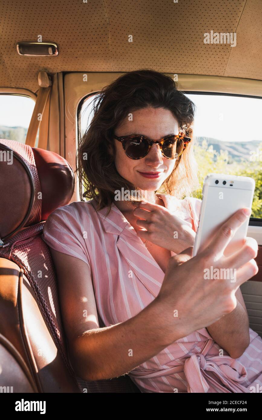 Schöne Brünette in rosa Kleid und trendige Sonnenbrille mit Telefon Und Selfie im Retro-Auto Stockfoto