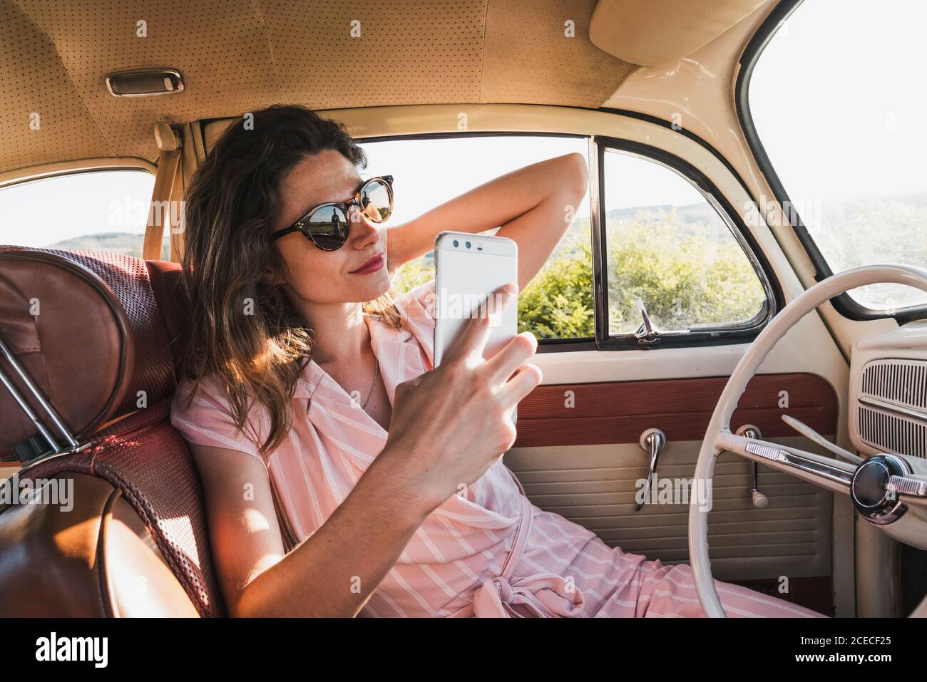 Schöne Brünette in rosa Kleid und trendige Sonnenbrille mit Telefon Und Selfie im Retro-Auto Stockfoto