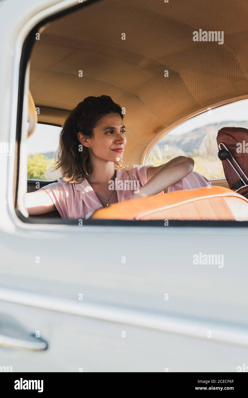 Schöne selbstbewusste Frau in Pink und Sonnenbrille auf dem Rücksitz im Retro-Auto sitzen und im Sonnenlicht wegschauen Stockfoto