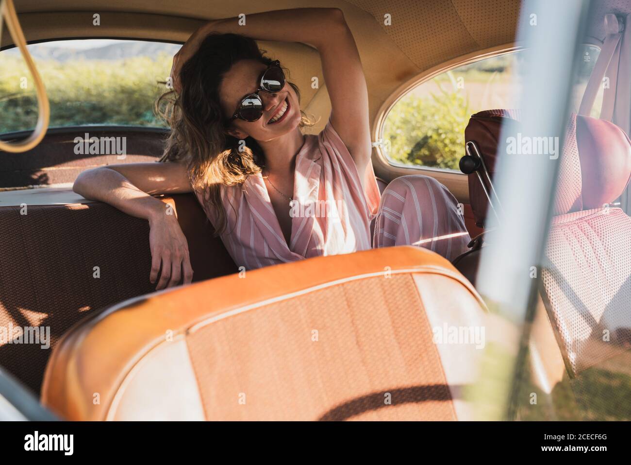 Schöne selbstbewusste Frau in Pink und Sonnenbrille auf dem Rücksitz im Retro-Auto sitzen und im Sonnenlicht wegschauen Stockfoto