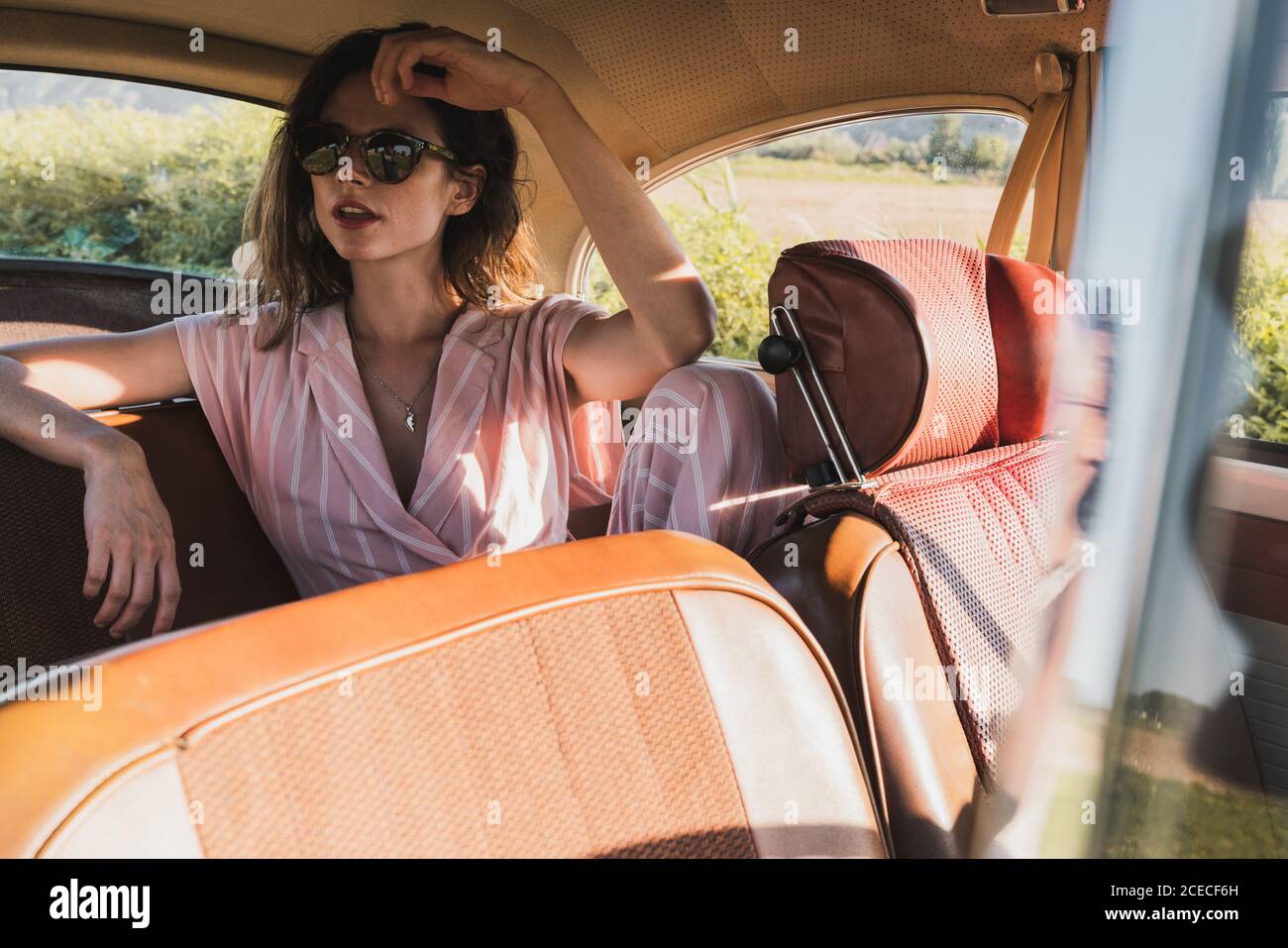 Schöne selbstbewusste Frau in Pink und Sonnenbrille auf dem Rücksitz im Retro-Auto sitzen und im Sonnenlicht wegschauen Stockfoto
