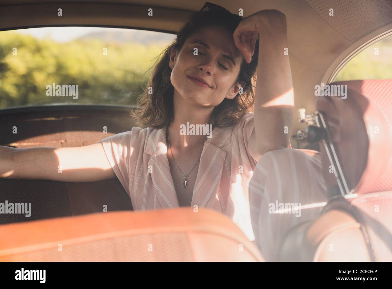 Schöne selbstbewusste Frau in Pink und Sonnenbrille auf dem Rücksitz im Retro-Auto sitzen und im Sonnenlicht wegschauen Stockfoto